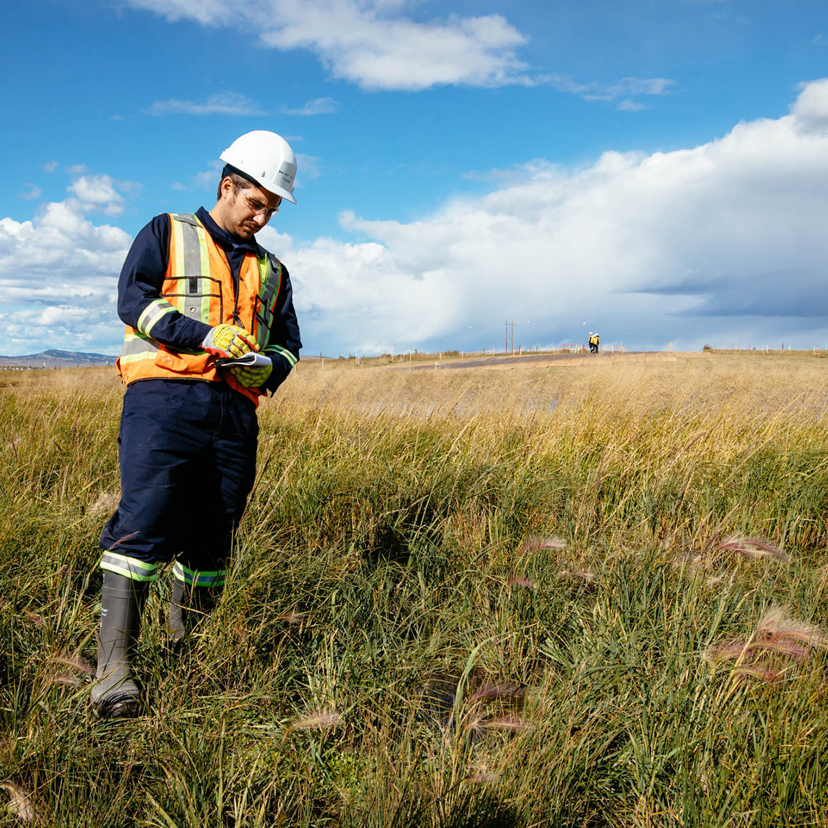 Person wearing personal protective equipment inspecting the ground in a field, notepad in hand.