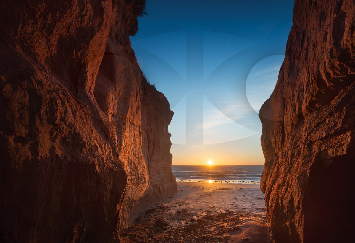 Sunrise over water between red rock cliffs on a beach in the Magdalen Islands, Quebec, featuring a transparent IAC logo.