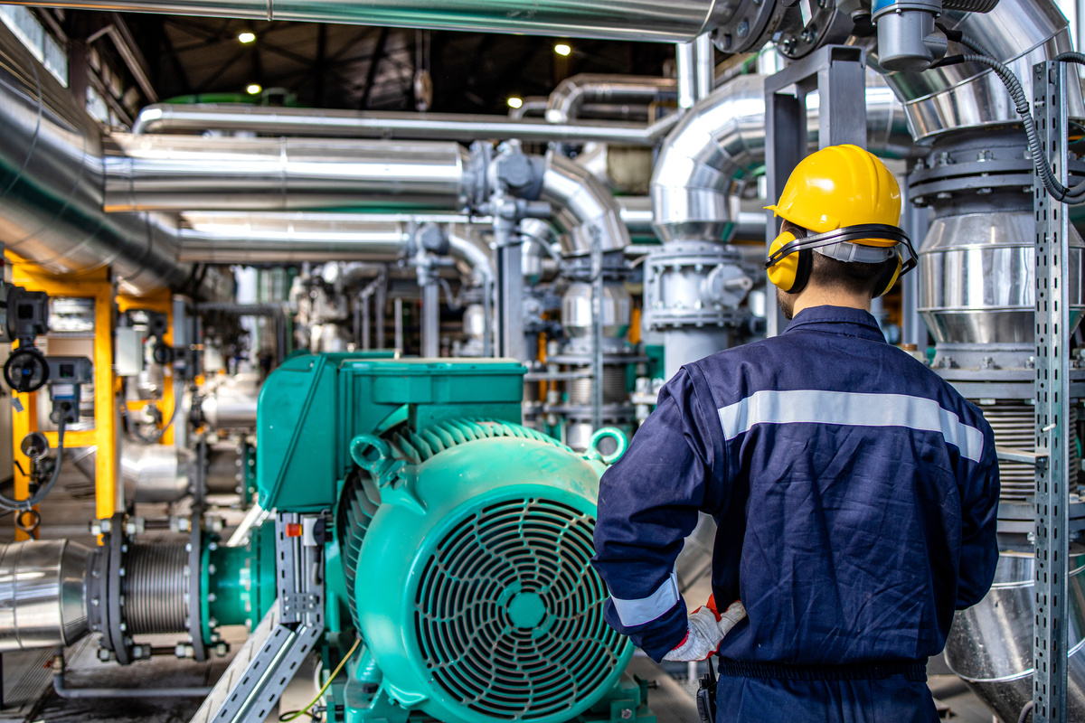Worker in PPE standing by gas fuel engines inside power plant checking production of electricity.
