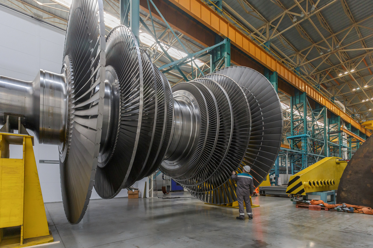 A worker in PPE assembles a large steam turbine rotor in a plant workshop.