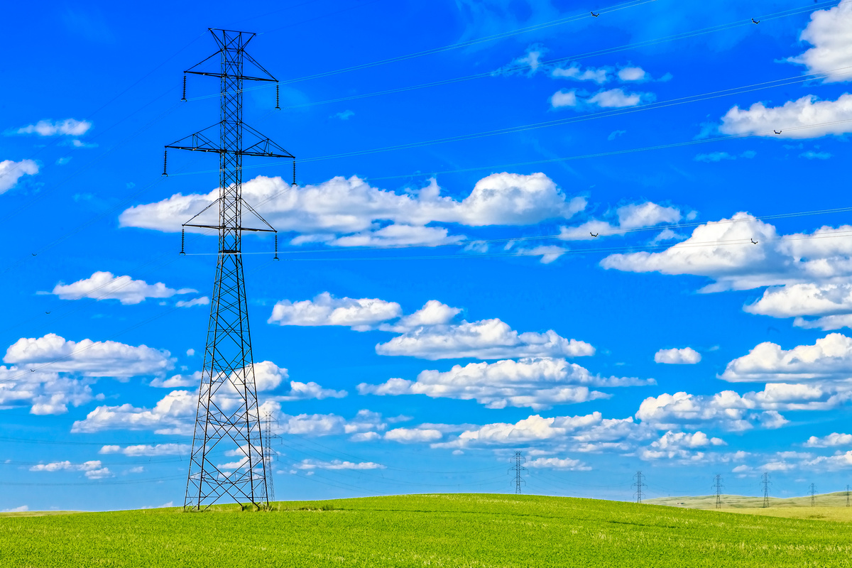 Electricity transmission towers linking powerlines traverse a green field. The sky is bright blue with fluffy white clouds.