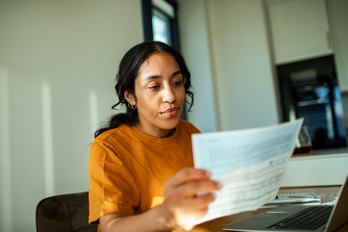 Young women in orange shirt sits at a desk in front of laptop computer reviewing a paper document, an electricity bill.