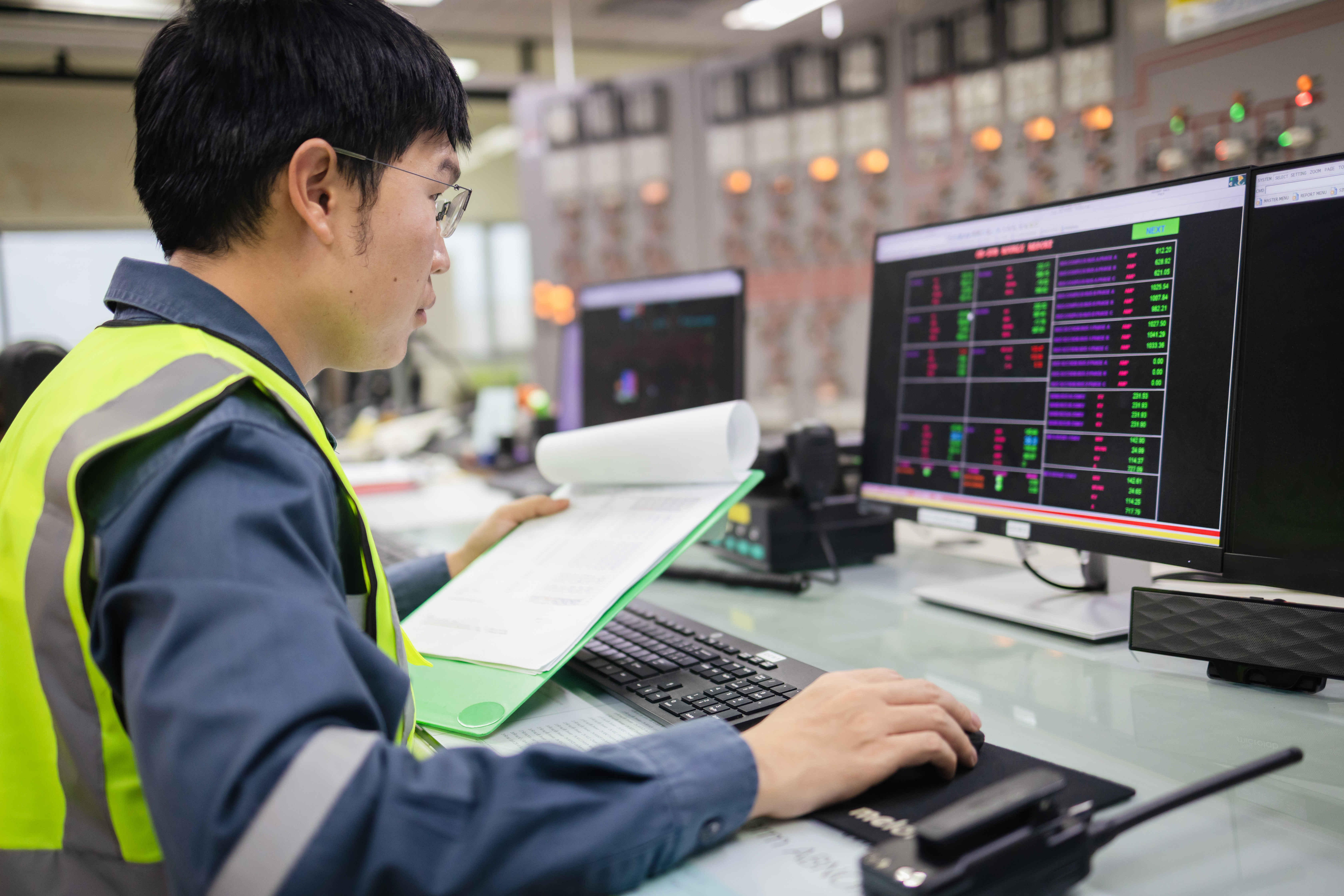Side view of an engineer in a safety vest monitoring electrical grid system on two monitors.