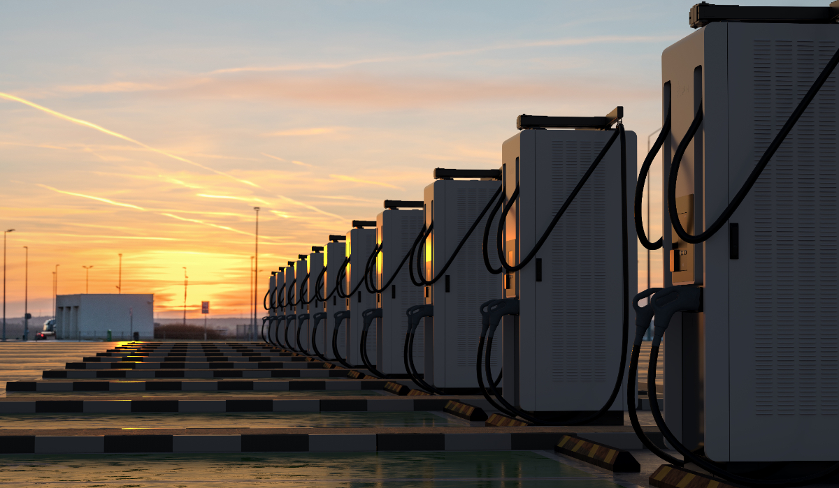 Several electric vehicle fast charging stations lined up in a parking lot with the sun setting behind them.