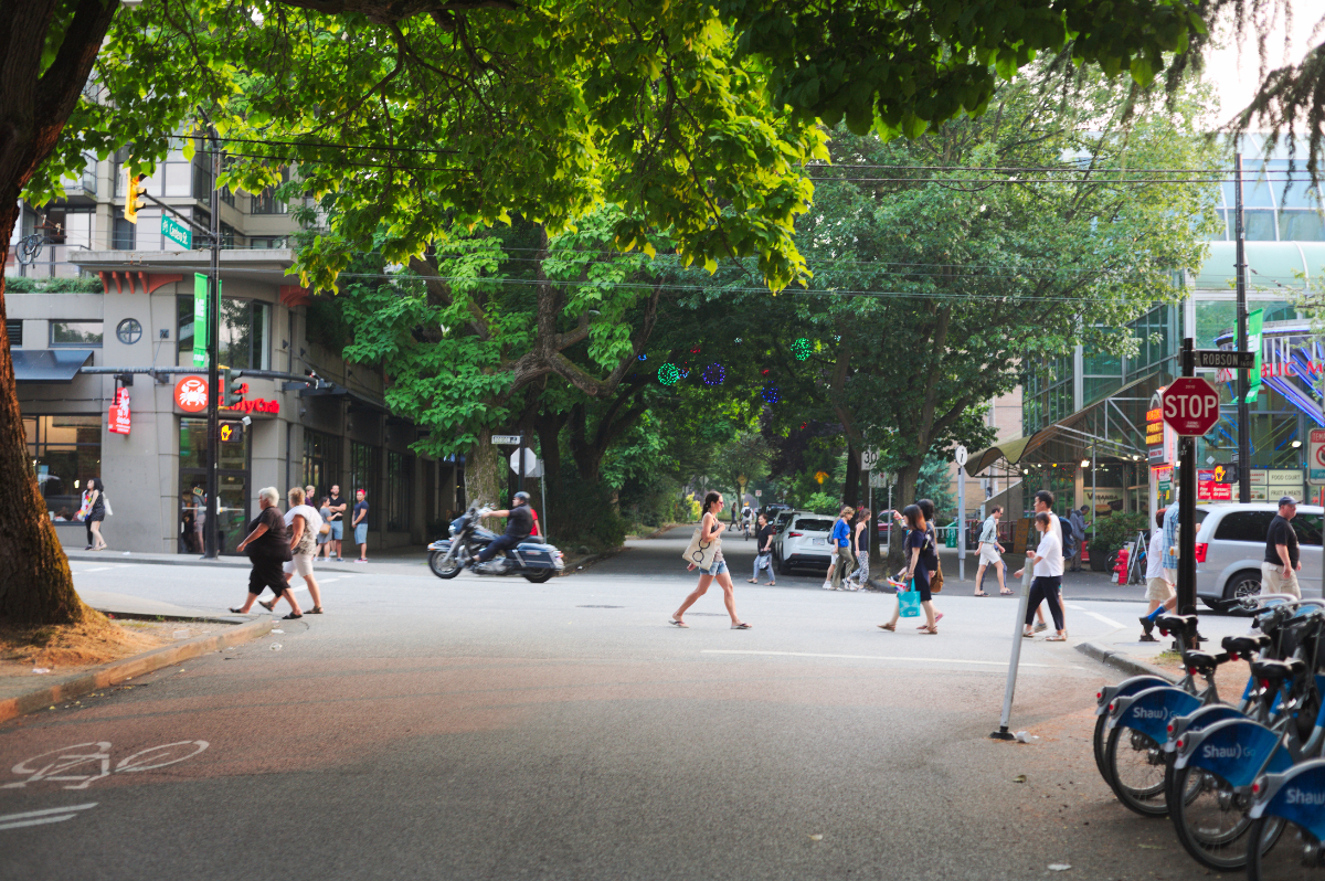 Busy city street in downtown Vancouver on a summer evening, pedestrians walking across the street and rental bikes are parked by a corner.