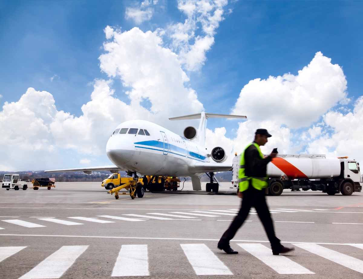 Airplane at a terminal, being serviced and fueled. Worker in visibility vest crosses foreground of image and fuel truck in seen in the distance.