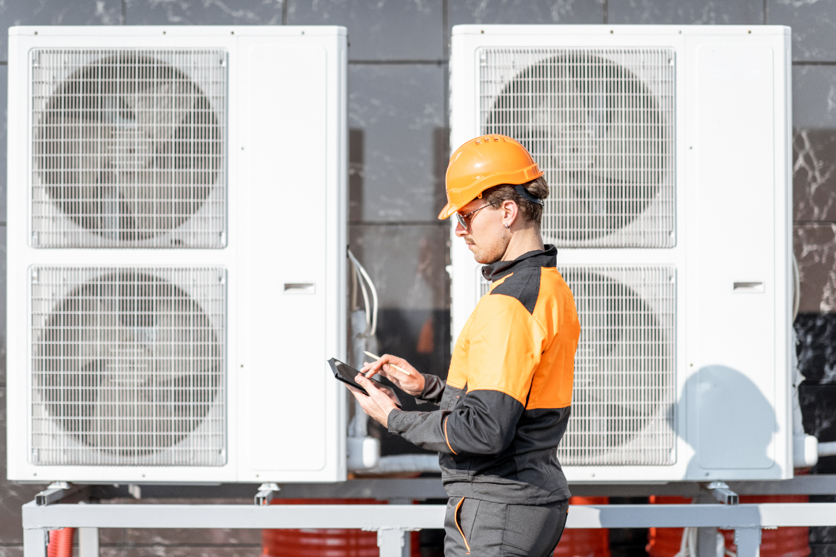 Man wearing orange PPE with tablet in hand, standing in front of two white heat pumps.