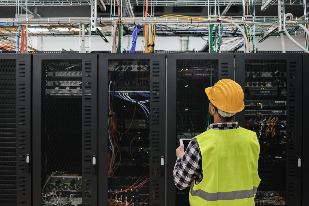 Young male technician in yellow helmet and vest, working with tablet inside big data center room full of rack servers.