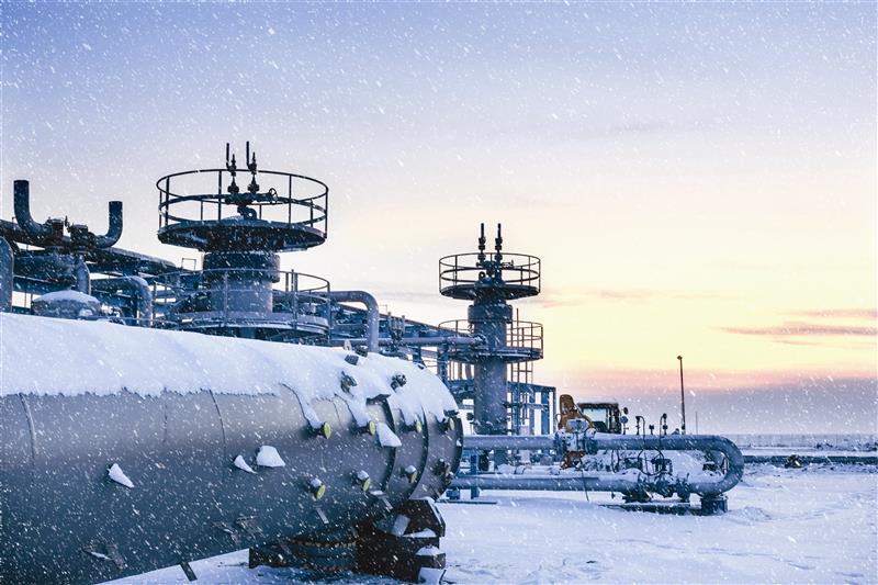 Station de compression de gaz naturel avec des valves et des tuyaux recouverts de neige lors d’une journée d’hiver froide. Le soleil se lève et il y a quelques nuages en arrière-plan.