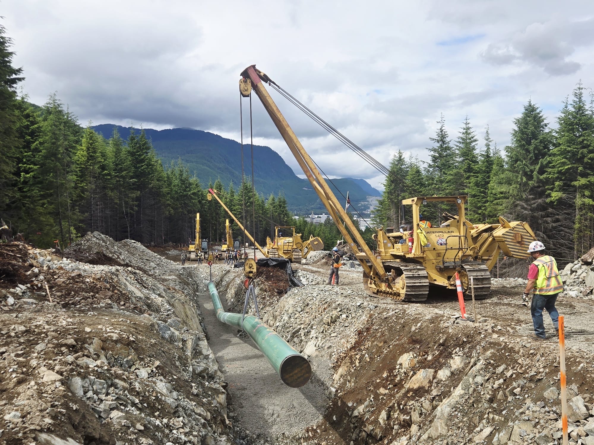 Des grues jaunes descendent le premier tuyau d’un pipeline de gaz naturel dans une tranchée, dans le nord de la Colombie-Britannique, alors que des travailleurs en équipement de protection individuelle supervisent le travail de long de la tranchée.