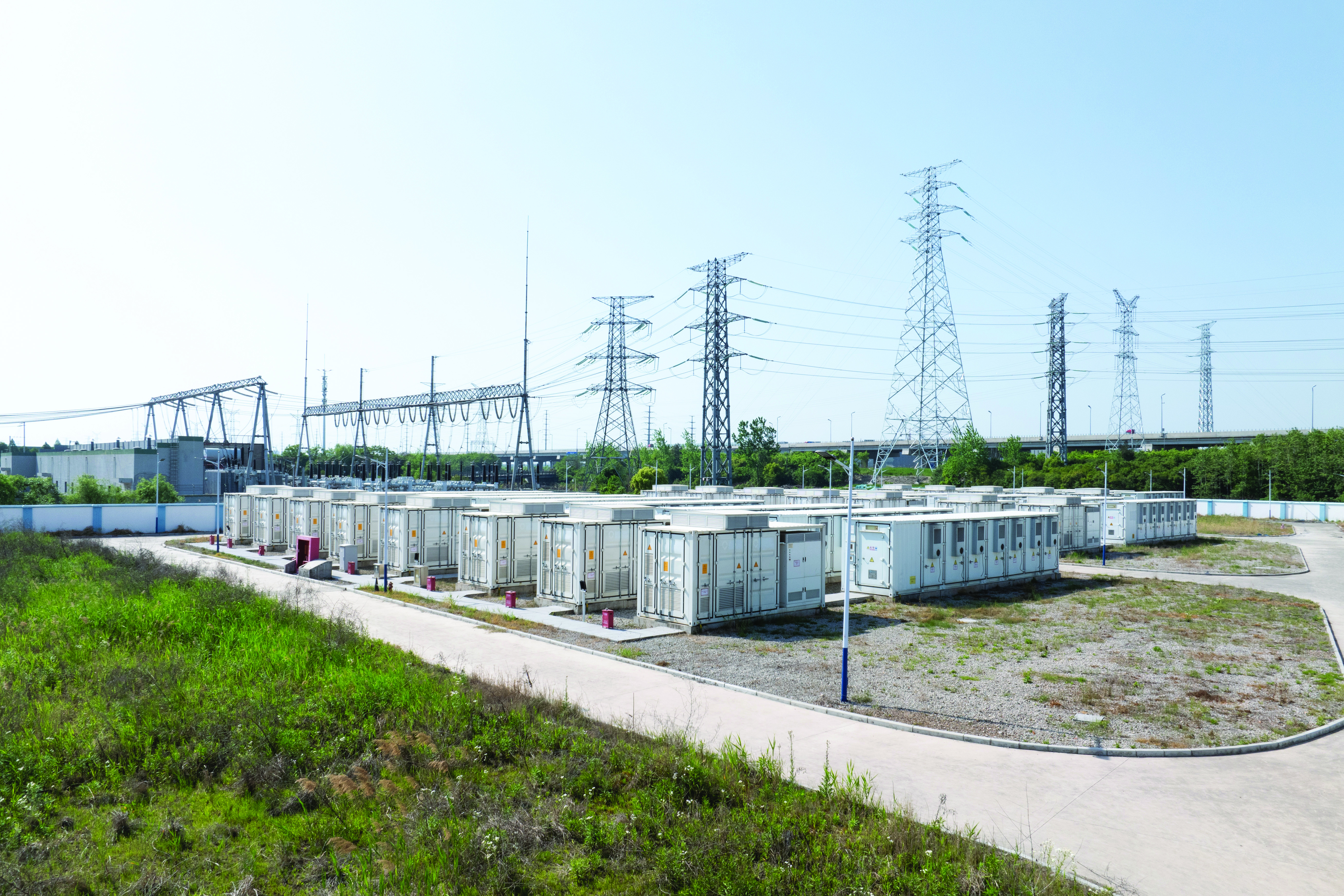 Batteries de stockage d’énergie blanches et grises sur un sol de gravier, près d’une sous-station, sous un ciel nuageux.